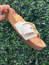 White platform slide sandal with a gold buckle held against a green leafy background