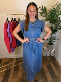 A woman modeling a sleeveless blue denim midi dress with a button-down front and a matching belt, standing in a boutique setting with a clothing rack and a potted plant in the background.