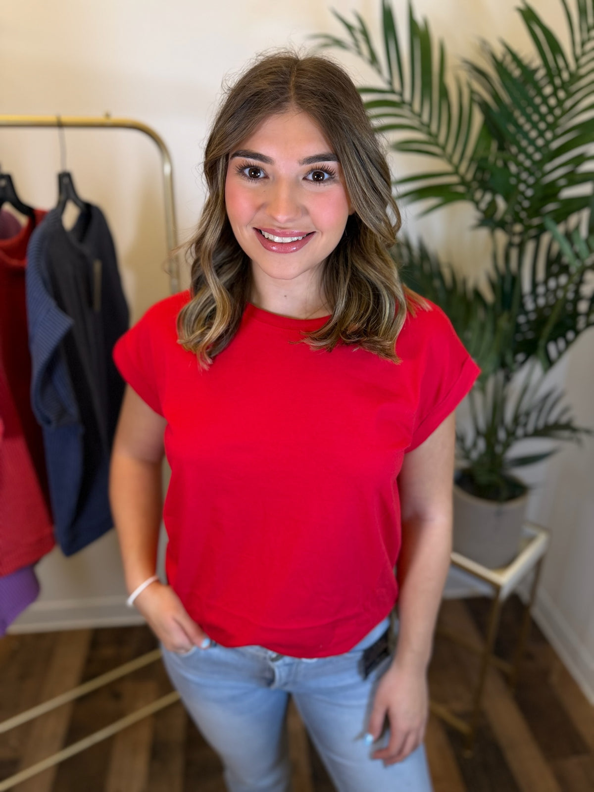 A woman smiling in a bright red short-sleeve t-shirt and light blue jeans, standing in a boutique setting with a clothing rack and a potted plant in the background.