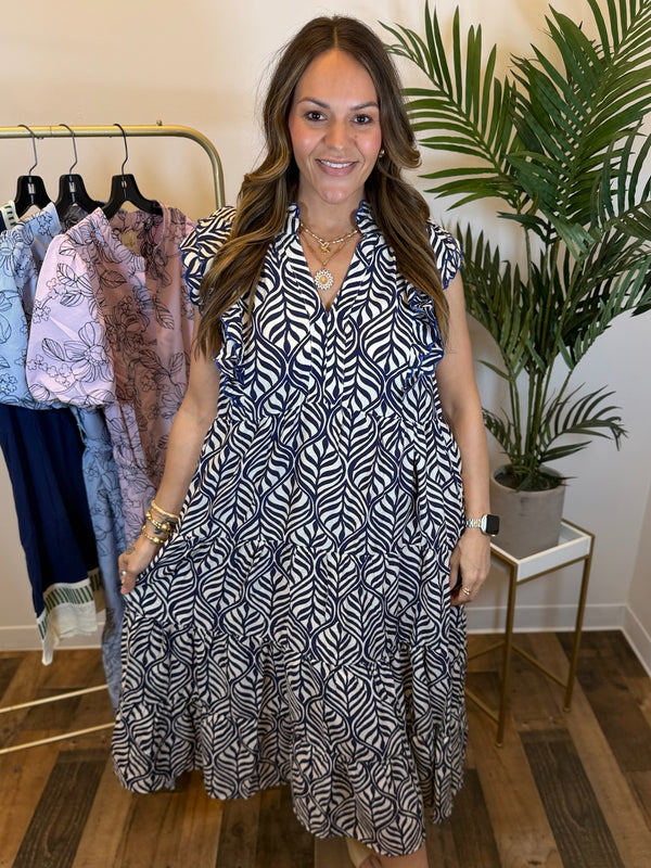Woman wearing a cream and navy patterned dress with scalloped trim and split neckline standing in a clothing store with plants and racks of clothes in the background.