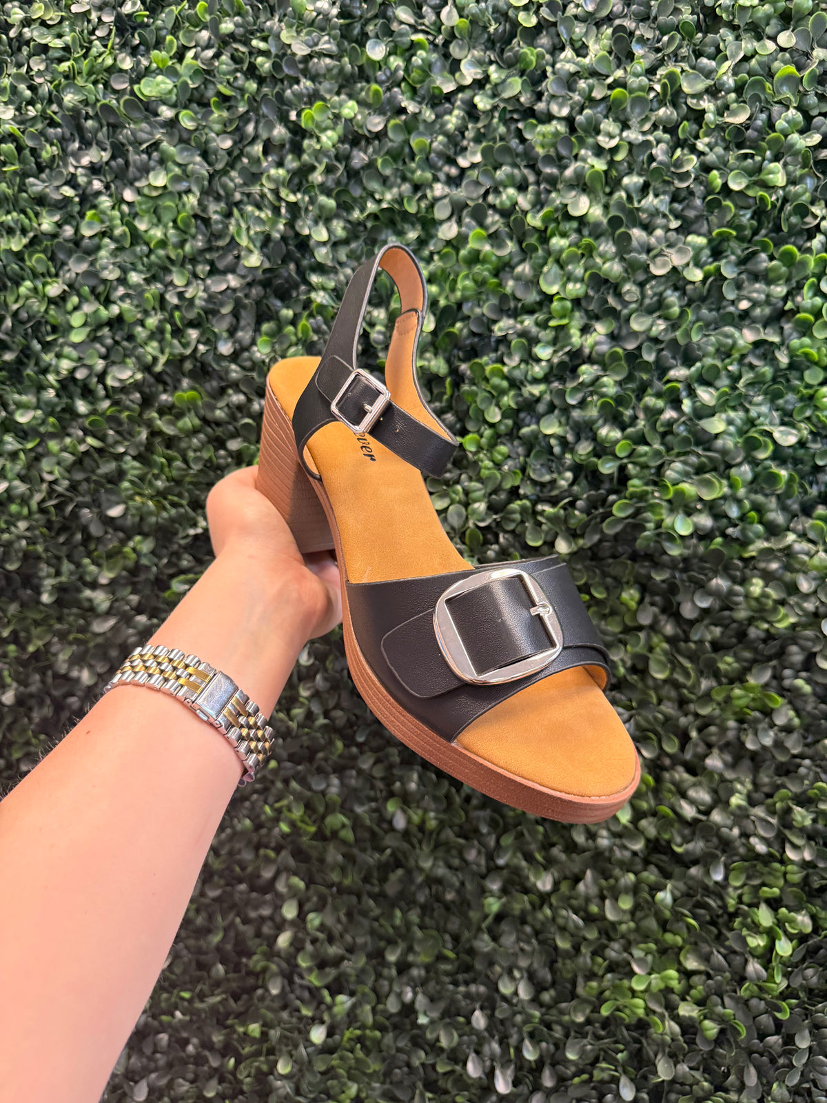 Hand holding a tan block heel with black straps against a green textured background