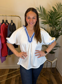 A woman modeling a white puff sleeve blouse with blue and white striped trim at the V-neck and cuffs, paired with dark blue jeans in an indoor boutique setting.