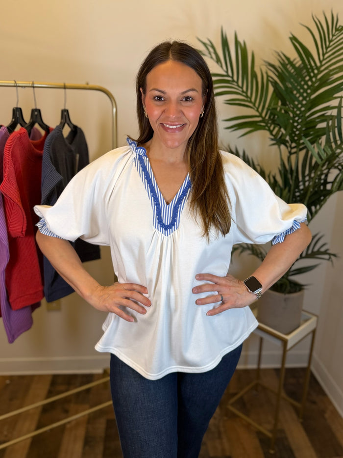 A woman modeling a white puff sleeve blouse with blue and white striped trim at the V-neck and cuffs, paired with dark blue jeans in an indoor boutique setting.