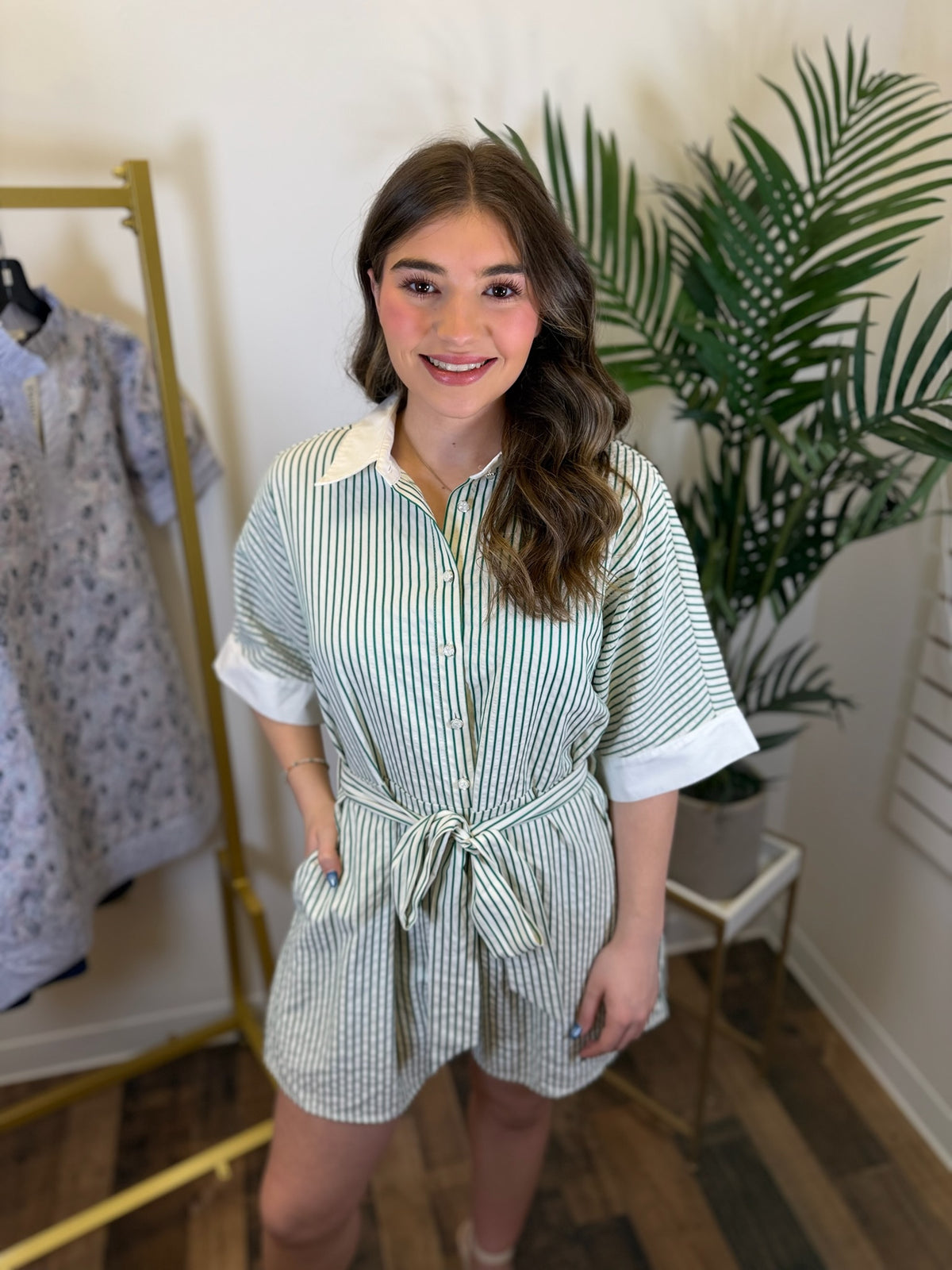 Woman wearing a green and white striped dress in a room with a plant and clothing rack.