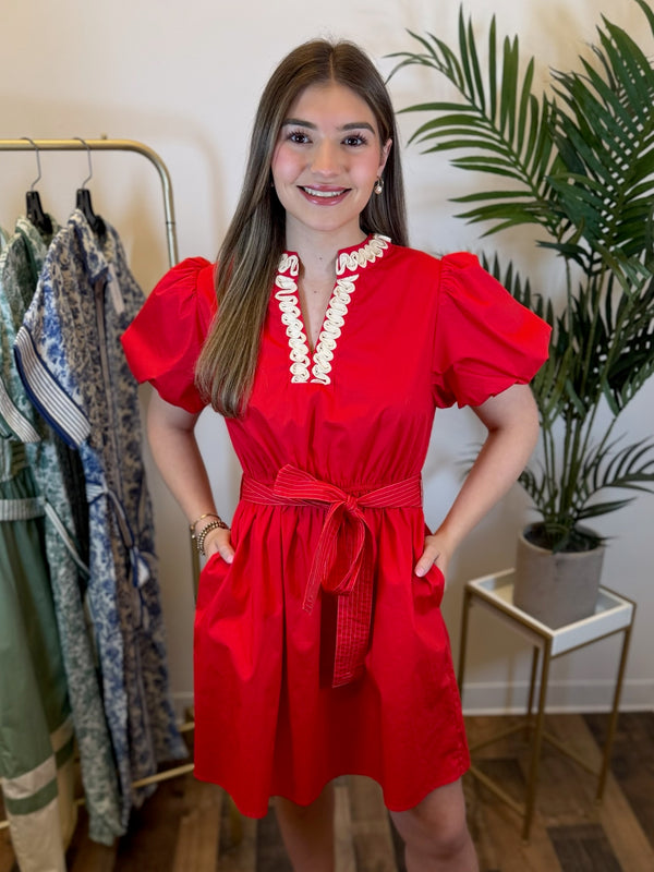 Woman wearing a Red mini dress with short puff sleeves and embroidered split neckline detail standing in a clothing store setting.