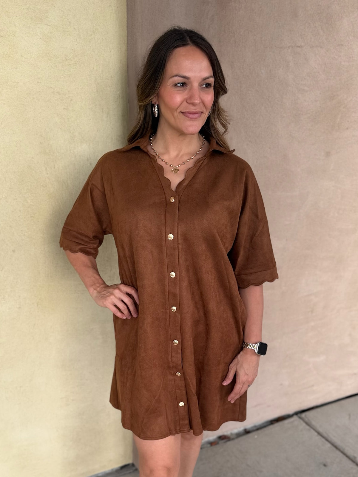 A woman wearing a short-sleeve camel button-front shirt dress with scalloped trim and pockets, standing outdoors against a beige stucco wall.