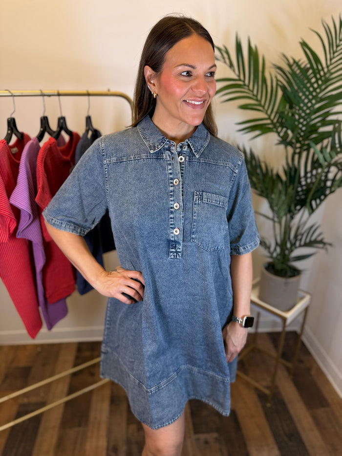 A woman wearing a short-sleeve denim shirt dress stands in a boutique setting with a clothing rack and a potted plant in the background.