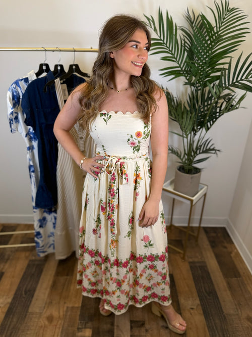 Woman wearing a cream floral midi dress with scalloped trim standing in a room with a rack of clothes and a plant.