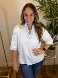 Woman wearing a white blouse with a collar, split neckline, and contrast stitch details in a room with a plant in the background