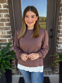 A woman wearing a mocha long-sleeve top with a white ruffled hem, paired with blue jeans, standing in front of a brick door.