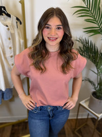 Woman wearing a pink top and blue jeans standing in a room with a plant and clothing rack.