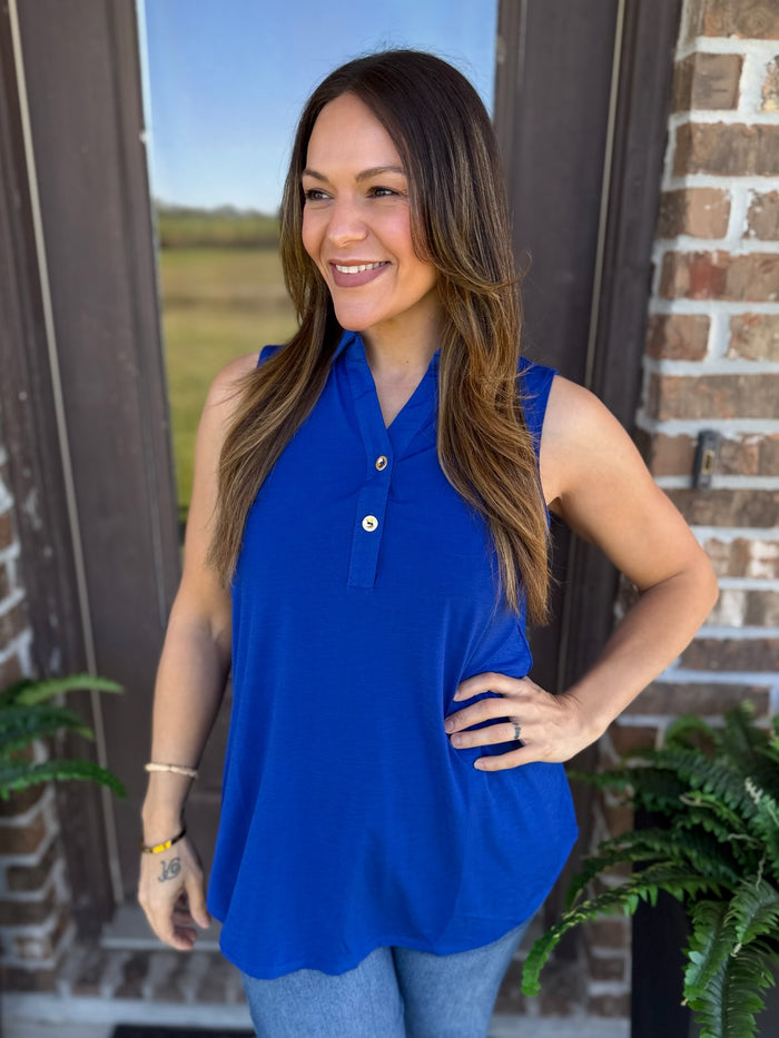 A woman wearing a sleeveless royal blue polo-style top with a button placket, standing outdoors in front of a brick wall and door frame.