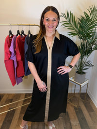 A woman modeling a black midi dress with a beige vertical placket and collar, standing in a boutique setting with a clothing rack and a potted plant in the background.