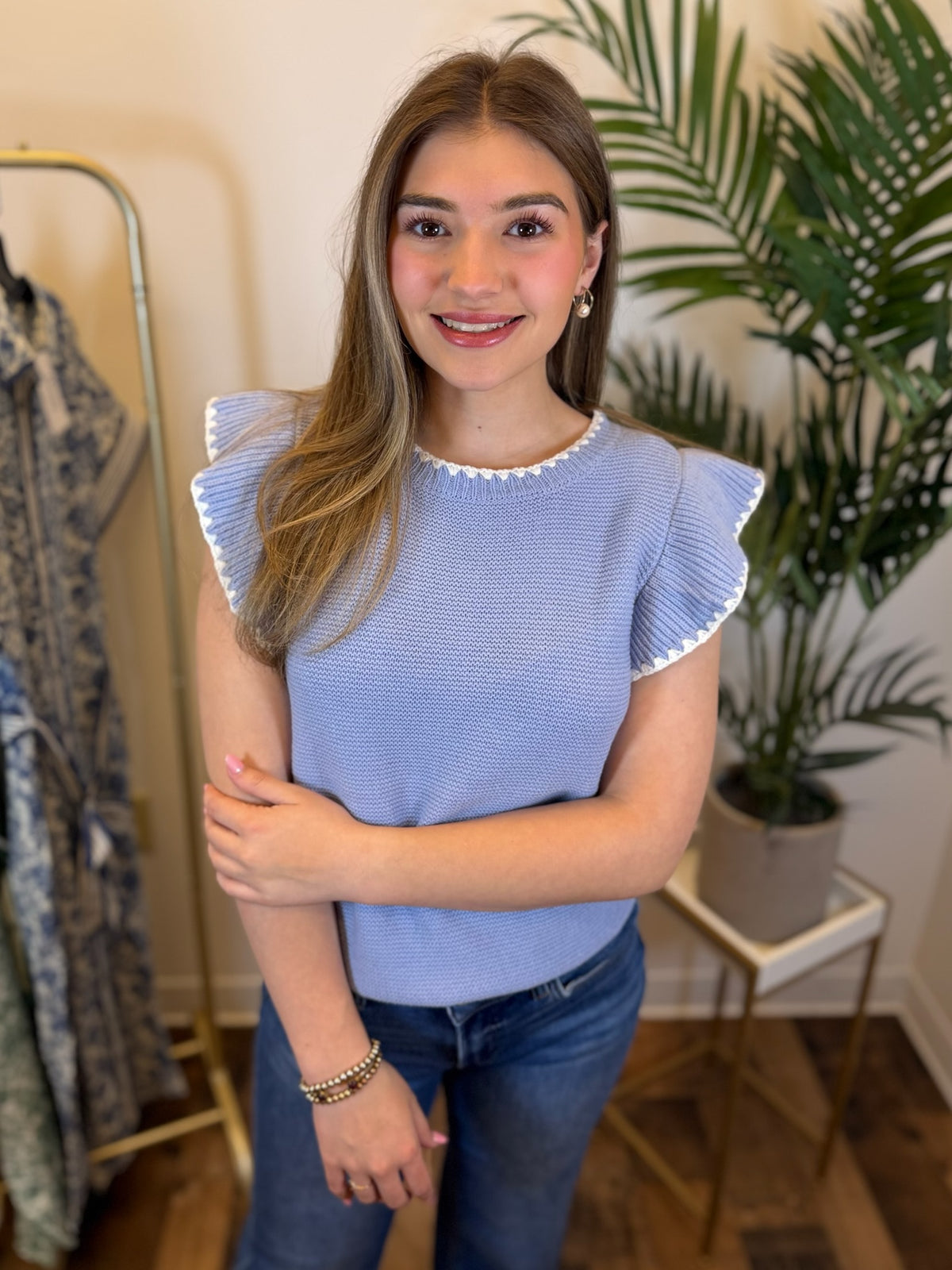 Woman wearing a blue top with ruffled sleeves and white trim details in a room with plants and a mirror.