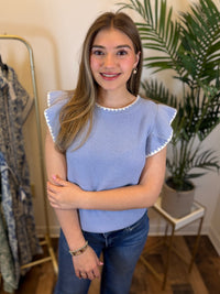 Woman wearing a blue top with ruffled sleeves and white trim details in a room with plants and a mirror.