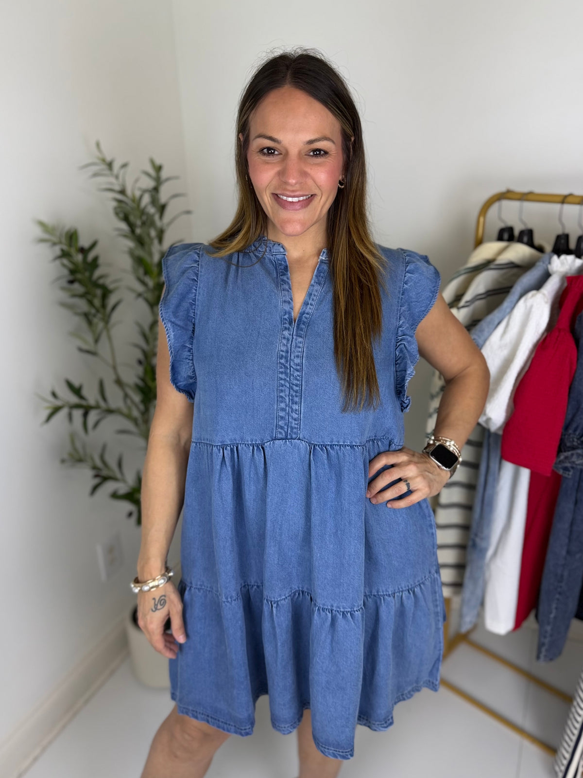 Woman wearing a tiered blue jean mini dress with split neckline and flutter cap sleeves standing in a room with a plant and clothing rack.