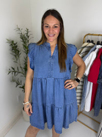 Woman wearing a tiered blue jean mini dress with split neckline and flutter cap sleeves standing in a room with a plant and clothing rack.