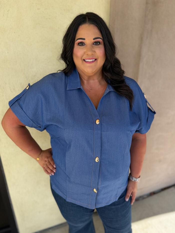 A woman wearing a blue short-sleeve button-up shirt with gold buttons and blue jeans, standing against a beige stucco wall.