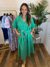 Woman in a kelly green midi dress with split neckline and contrast trim details standing in a clothing store with plants and racks of clothes in the background.