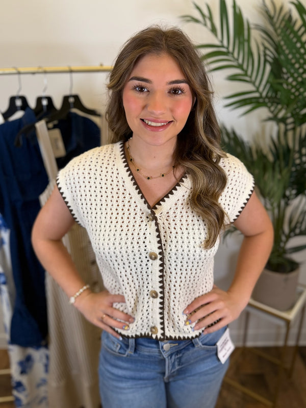 Woman wearing a white crochet top with brown buttons and embroidered trim in a clothing store.
