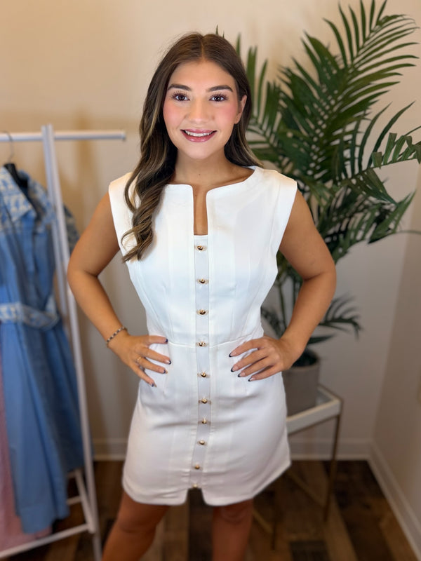 Woman wearing a white dress with gold buttons in a room with a plant and clothing rack.