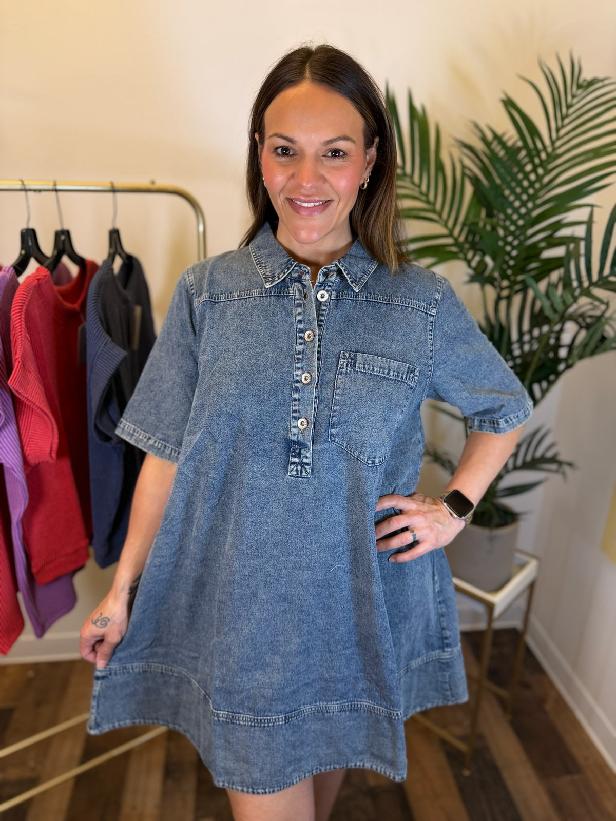 A woman wearing a short-sleeve blue denim shirt dress with a buttoned henley collar, standing in a boutique setting with a clothing rack and a potted plant in the background.
