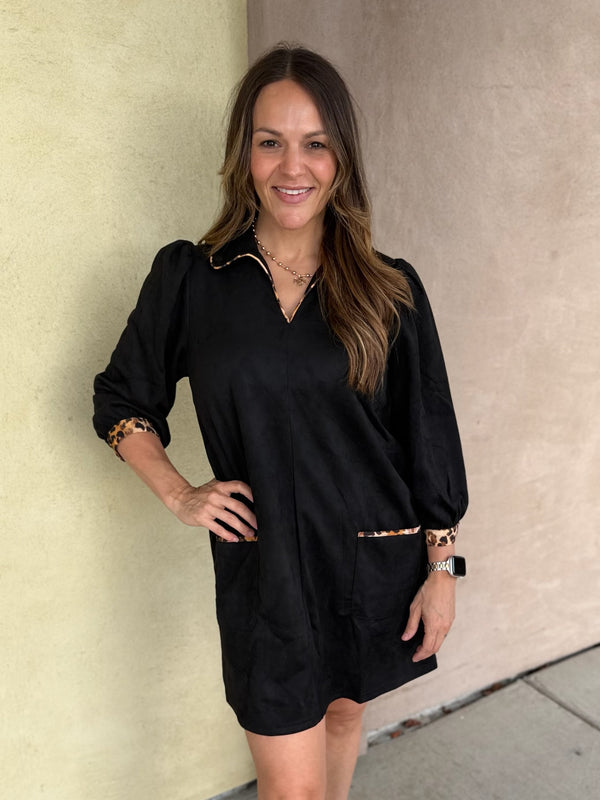 A woman modeling a black long-sleeve mini dress with collared v neck, leopard trim, and pockets, paired with white ankle boots, standing against a beige stucco wall.