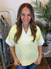 Woman wearing a light yellow blouse with white trim, short sleeves, and split neckline in a clothing store.