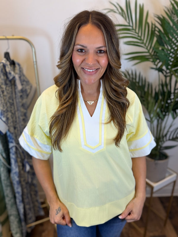 Woman wearing a light yellow blouse with white trim, short sleeves, and split neckline in a clothing store.