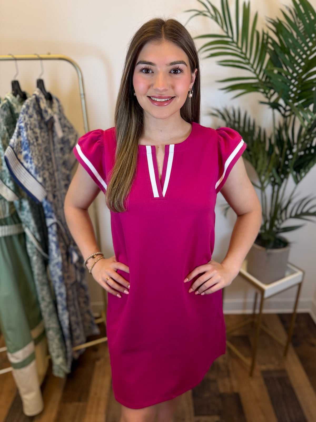 woman wearing a fuchsia v neck dress with ruffled cap sleeves and contrast trim details posing in a room with a plant and clothing rack.