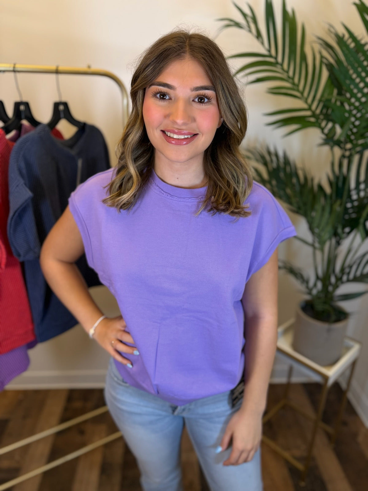 A woman wearing a lavender purple short-sleeve crewneck t-shirt and light blue jeans, standing in a boutique setting with a clothing rack and a potted plant in the background.