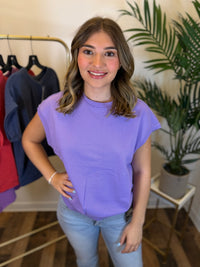 A woman wearing a lavender purple short-sleeve crewneck t-shirt and light blue jeans, standing in a boutique setting with a clothing rack and a potted plant in the background.