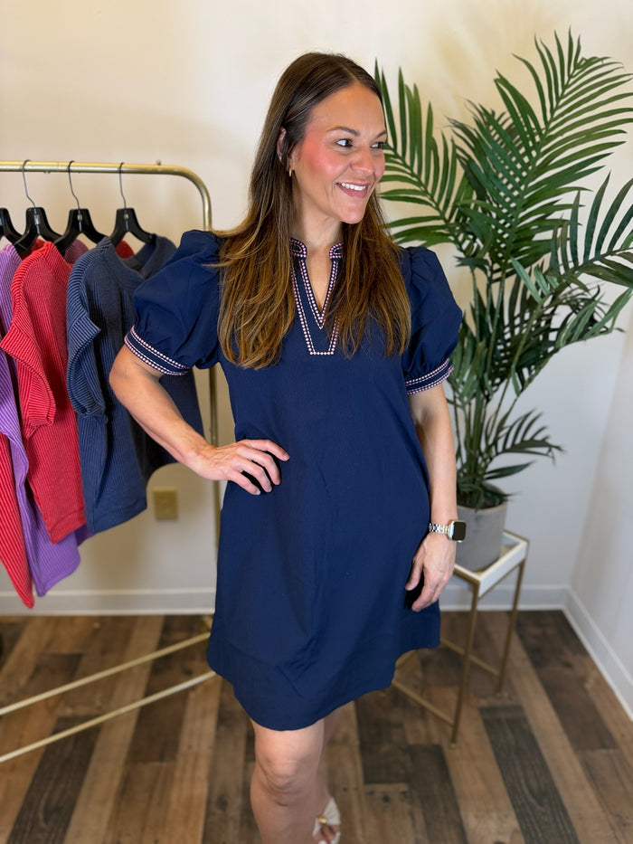 A woman modeling a navy blue puff-sleeve mini dress with a V-neckline and pink embroidered trim, standing in a boutique setting with a clothing rack and a potted plant in the background.
