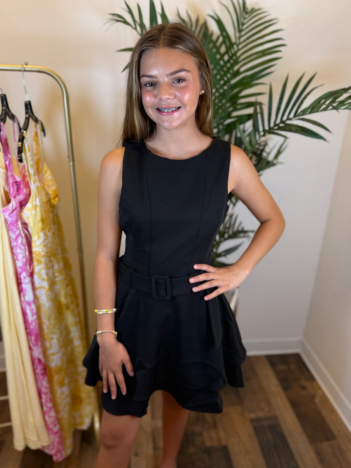 Young woman wearing a black sleeveless dress in a room with a plant and rack of dresses.