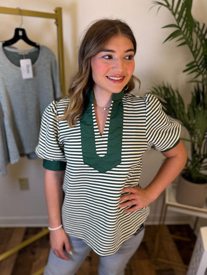 A woman modeling a short-sleeve top with green and white horizontal stripes and a solid green V-neck trim, standing in a boutique setting.