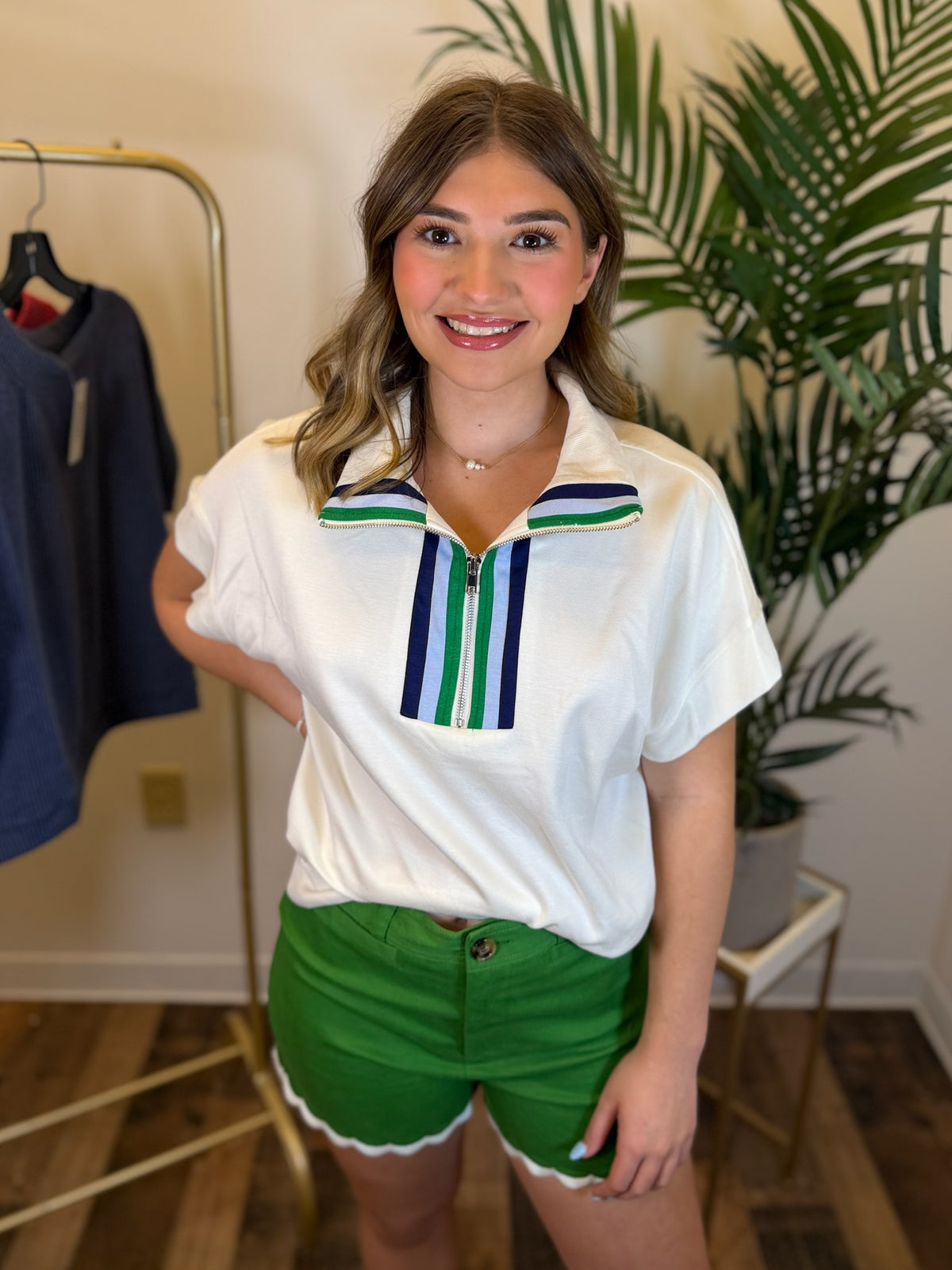A woman modeling a white short-sleeve polo-style top with green and navy striped collar details, paired with bright green shorts, standing in an indoor room with a clothing rack and potted plant in the background.