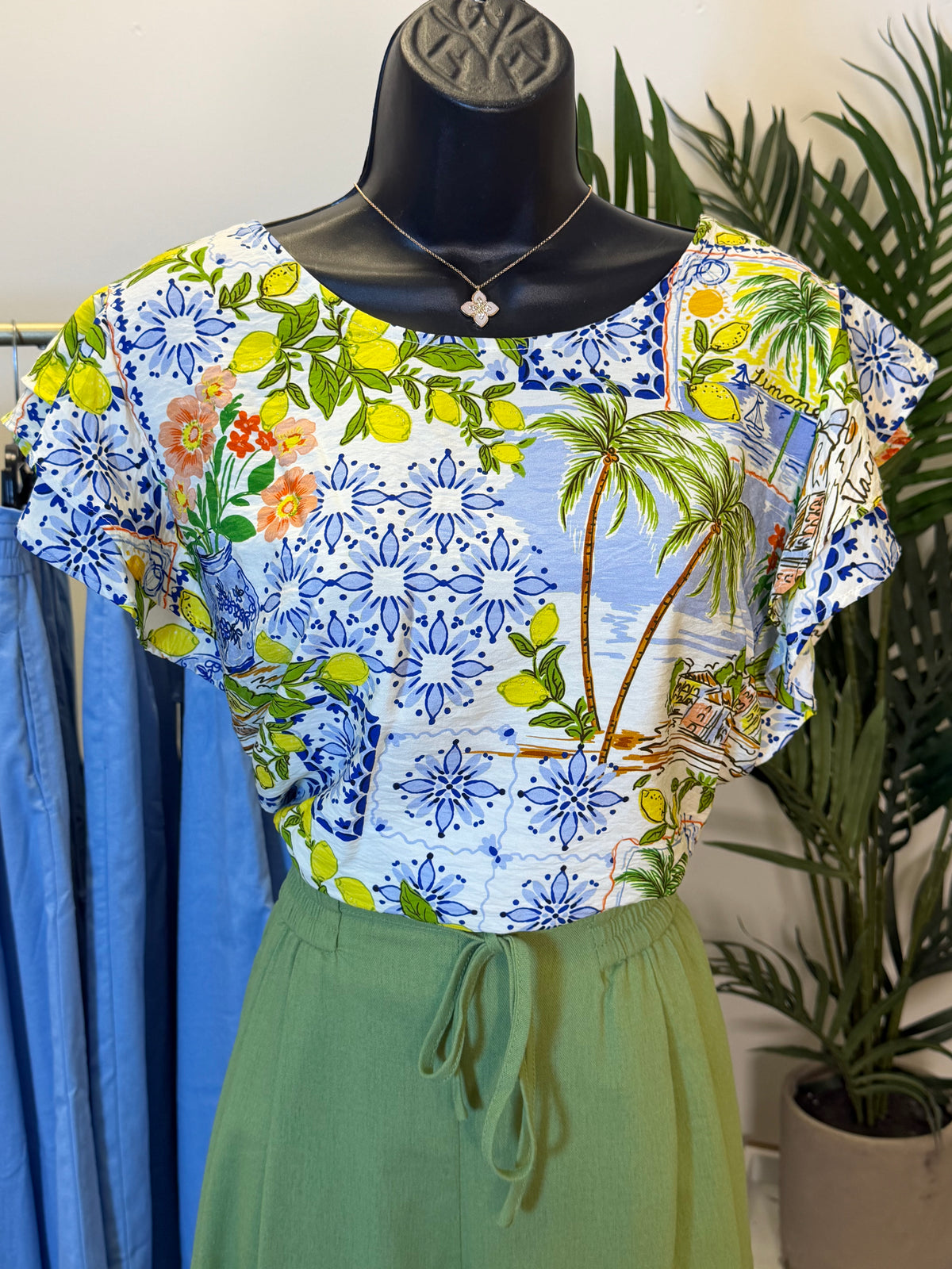 Colorful floral and palm tree patterned ruffle sleeve blouse on a mannequin with a plant in the background