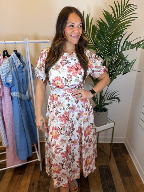 Woman wearing a cream dress with pink floral print and puff sleeves in a clothing store with plants and racks of clothes in the background.