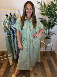 Woman wearing a green striped maxi dress with button front and pleated waist details standing in a room with a rack of clothes and a plant