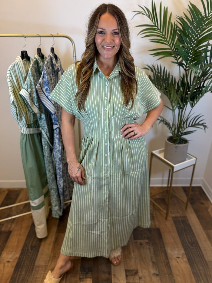 Woman wearing a green striped maxi dress with button front and pleated waist details standing in a room with a rack of clothes and a plant