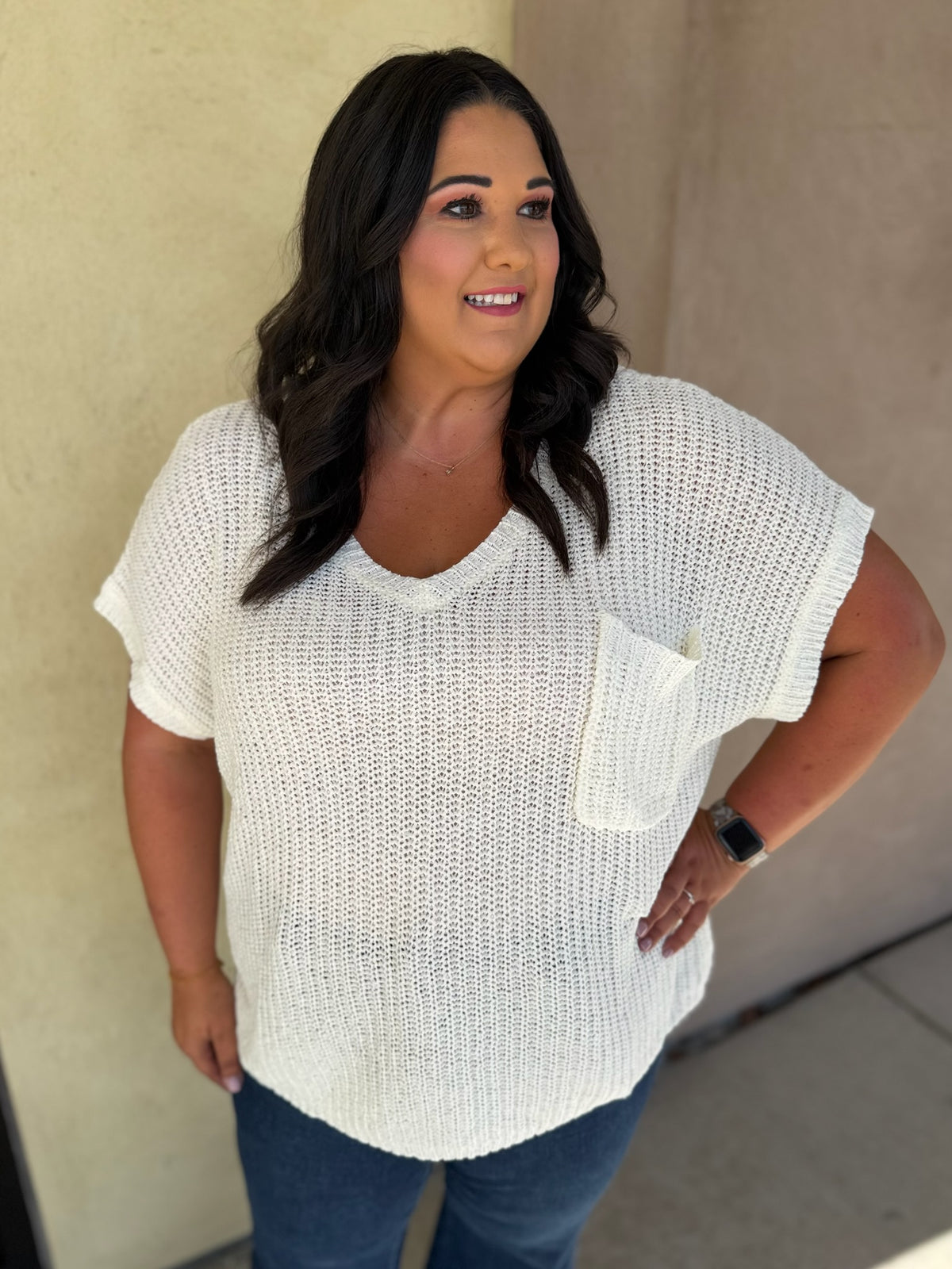 A woman wearing a white textured knit short-sleeve top with a chest pocket, paired with blue jeans, standing against a beige stucco wall.