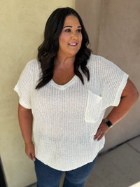 A woman wearing a white textured knit short-sleeve top with a chest pocket, paired with blue jeans, standing against a beige stucco wall.