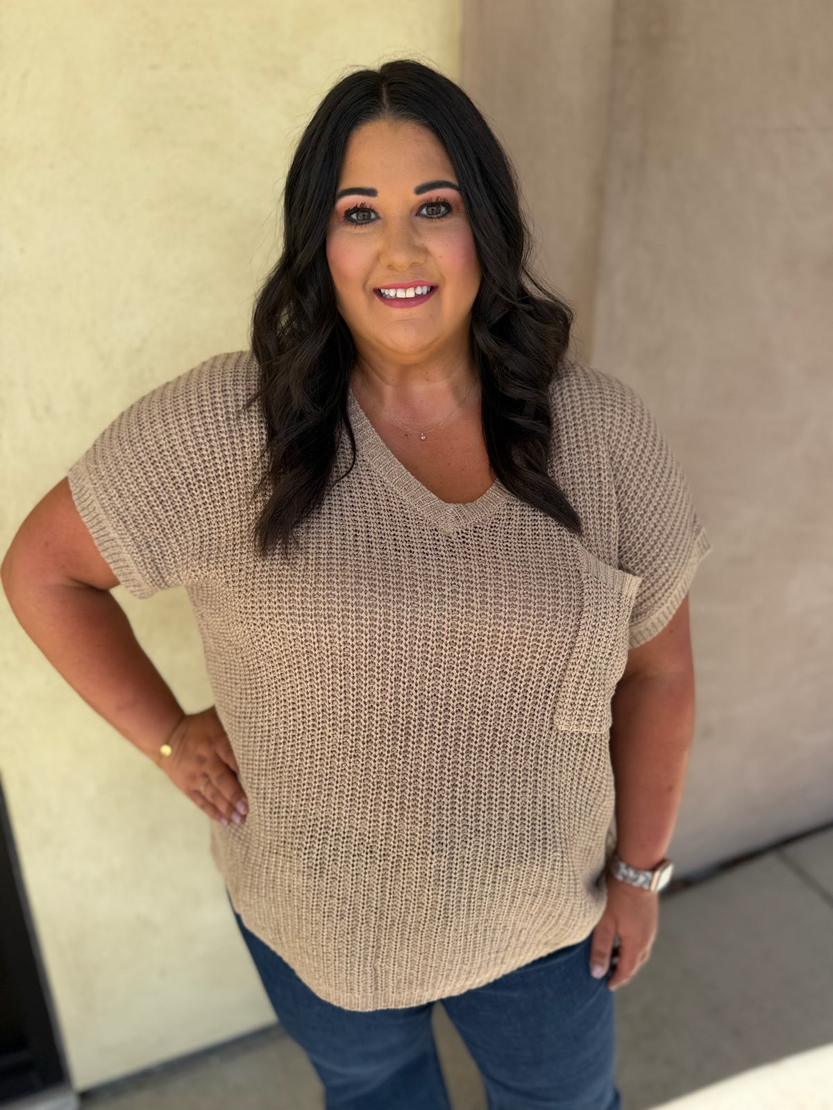 A woman wearing a beige short-sleeve knit V-neck top with a chest pocket, paired with blue jeans, standing against a neutral beige wall.