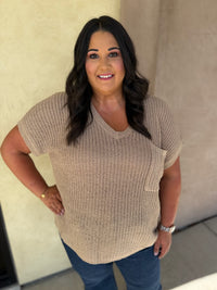 A woman wearing a beige short-sleeve knit V-neck top with a chest pocket, paired with blue jeans, standing against a neutral beige wall.