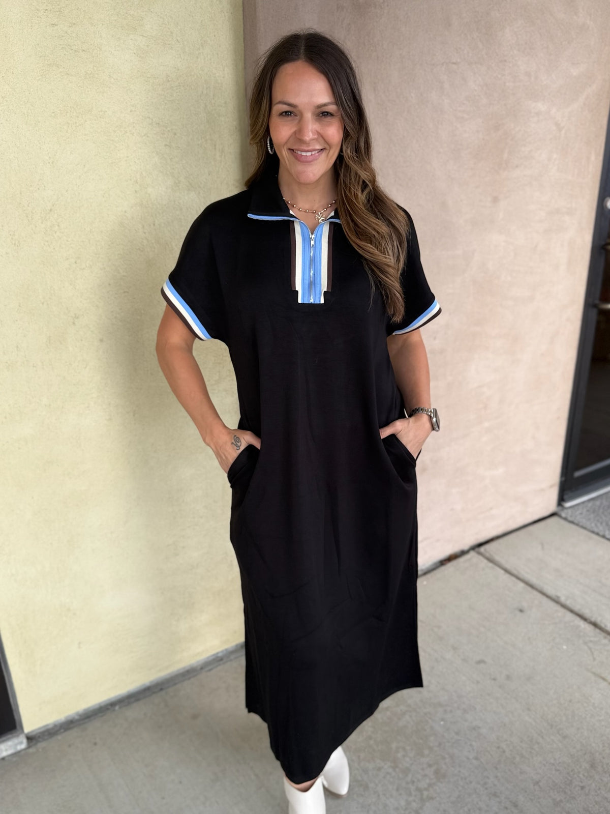 A woman modeling a black short-sleeve midi dress with light blue and white striped trim on the collar and sleeve cuffs, paired with white ankle boots, standing against a beige stucco wall.