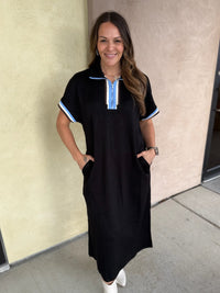A woman modeling a black short-sleeve midi dress with light blue and white striped trim on the collar and sleeve cuffs, paired with white ankle boots, standing against a beige stucco wall.