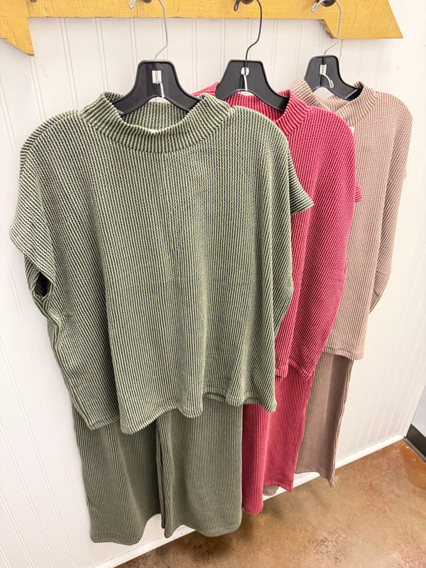 Four ribbed knit sleeveless crop tops in beige, burgundy, sage green, and olive green, displayed on black hangers on a white slatted surface.