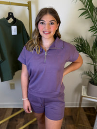 A woman modeling a matching purple lounge set consisting of a short-sleeve quarter-zip pullover and matching shorts, standing in a boutique setting with a clothing rack and plant in the background.