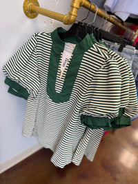 A short-sleeve top with green and off-white horizontal stripes, featuring a solid green mock neck and ruffled trim, hanging on a black hanger on a gold industrial pipe rack.