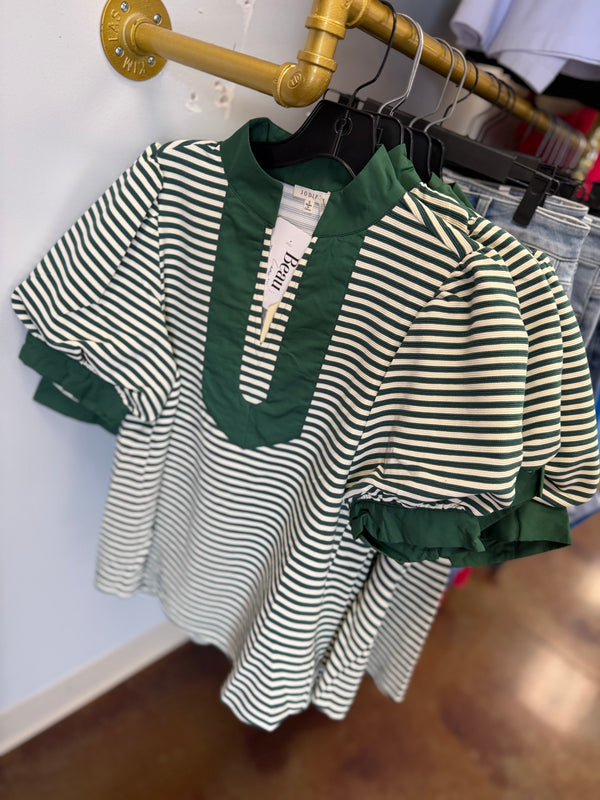 A short-sleeve top with green and off-white horizontal stripes, featuring a solid green mock neck and ruffled trim, hanging on a black hanger on a gold industrial pipe rack.